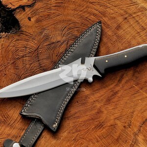 May include: A stainless steel dagger with a black handle and a leather sheath. The dagger has a double-edged blade and a decorative guard. The sheath is black with white stitching. The dagger is resting on a wooden surface.