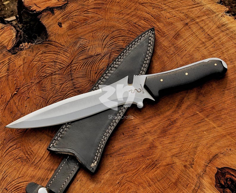 May include: A stainless steel dagger with a black handle and a leather sheath. The dagger has a double-edged blade and a decorative guard. The sheath is black with white stitching. The dagger is resting on a wooden surface.