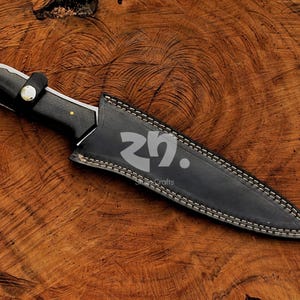 May include: A black hunting knife with a leather sheath, on a wooden surface. The knife has a black handle with brass rivets and a silver blade. The sheath is black, with white stitching and a strap.