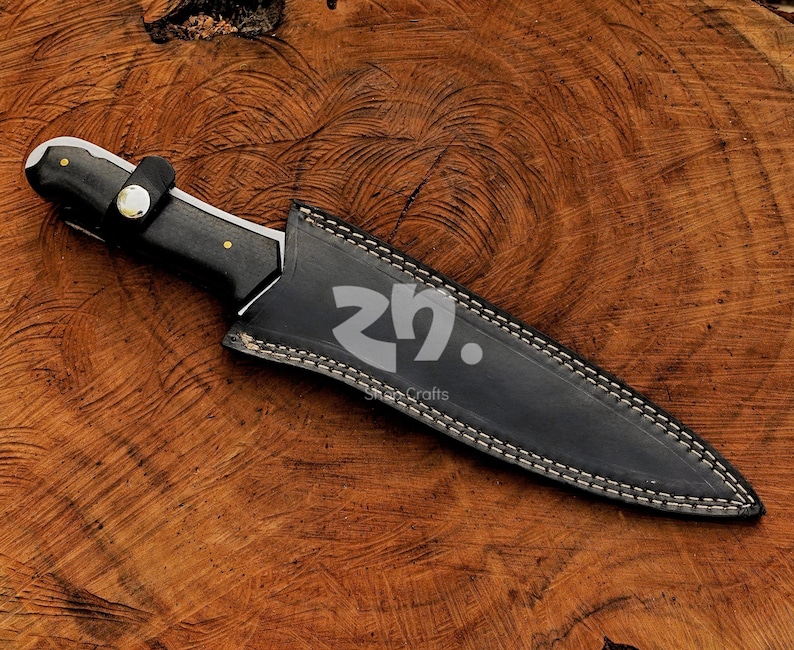 May include: A black hunting knife with a leather sheath, on a wooden surface. The knife has a black handle with brass rivets and a silver blade. The sheath is black, with white stitching and a strap.