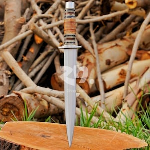 May include: A double-edged dagger with a pointed steel blade and a cylindrical handle featuring alternating bands of light brown wood and dark brown rings, finished with a metallic pommel and a small crossguard at the base of the handle.