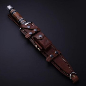 May include: A fixed-blade knife with a stacked leather handle featuring black and silver rings, housed in a dark brown leather sheath. The sheath includes a small front pouch, multiple metal snap closures, visible stitching, and a metal D-ring.