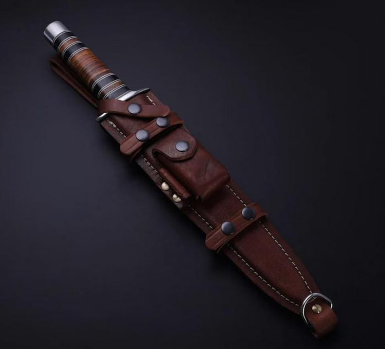 May include: A fixed-blade knife with a stacked leather handle featuring black and silver rings, housed in a dark brown leather sheath. The sheath includes a small front pouch, multiple metal snap closures, visible stitching, and a metal D-ring.