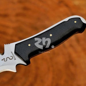 May include: Close-up of a knife with a black handle and a silver blade. The handle has three gold-coloured rivets. The blade features the text "Krauser" and a snake symbol. The knife is set against a wooden background.