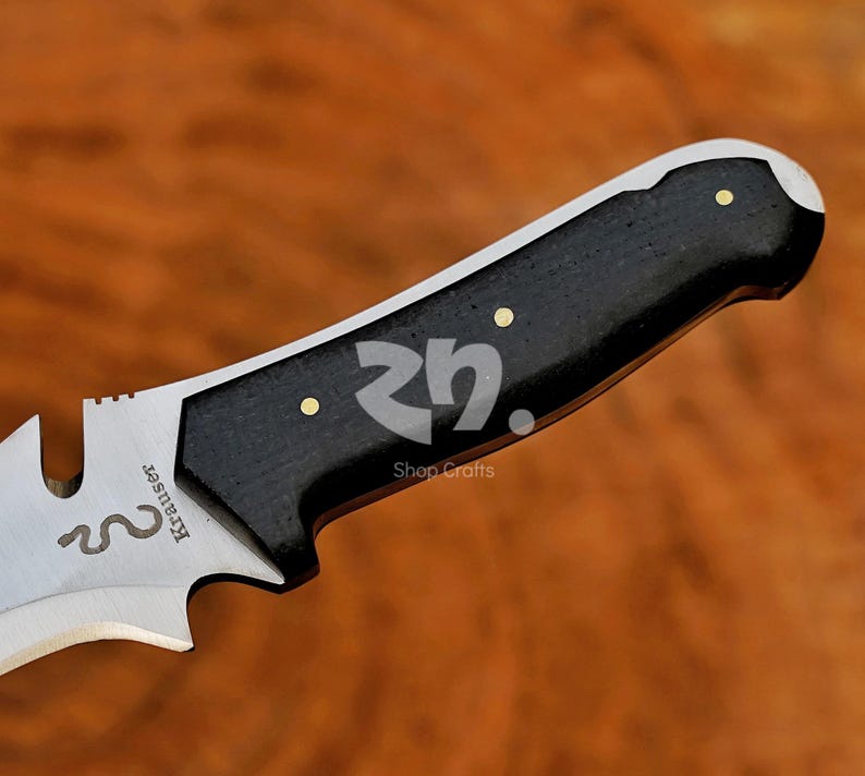 May include: Close-up of a knife with a black handle and a silver blade. The handle has three gold-coloured rivets. The blade features the text "Krauser" and a snake symbol. The knife is set against a wooden background.