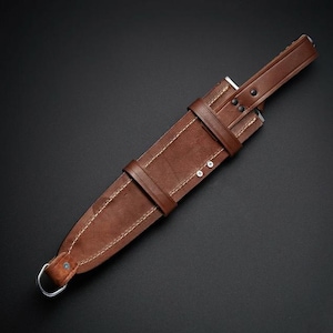 May include: A brown leather sheath featuring visible light-colored stitching, a secure strap with two metal rivets, and a metal D-ring at the bottom. The sheath is designed with a tapered shape and includes a reinforced top section with additional rivets.