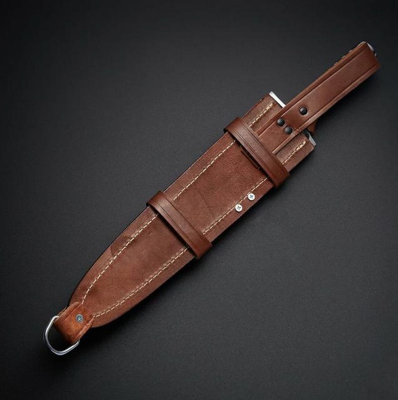 May include: A brown leather sheath featuring visible light-colored stitching, a secure strap with two metal rivets, and a metal D-ring at the bottom. The sheath is designed with a tapered shape and includes a reinforced top section with additional rivets.