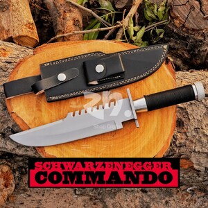 May include: A large, silver-bladed knife with a black handle and a black leather sheath. The knife has a serrated edge and a silver crossguard. The text "SCHWARZENEGGER COMMANDO" is displayed in a red and black banner.