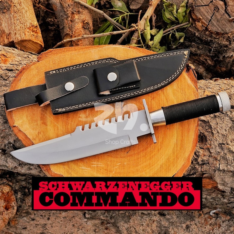 May include: A large, silver-bladed knife with a black handle and a black leather sheath. The knife has a serrated edge and a silver crossguard. The text "SCHWARZENEGGER COMMANDO" is displayed in a red and black banner.