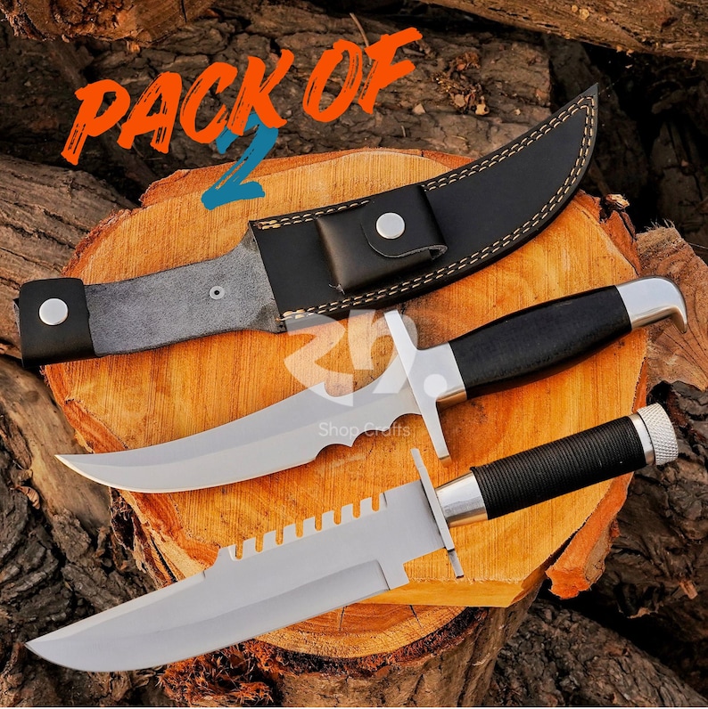May include: A pack of two hunting knives with silver blades and black handles. One knife has a serrated edge, and both have a silver crossguard. The knives rest on a wooden surface, with a black leather sheath.