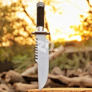 May include: A large, silver survival knife with a serrated edge and a black, textured handle. The knife has a silver guard and a silver cap on the handle. The text "Shop Crafts" is visible on the blade.