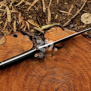 May include: A black-handled knife with a silver blade rests on a textured, brown wooden surface. The knife's blade is sharp and angled, with a visible edge. The handle has a silver stripe. The background includes wood shavings and leaves.