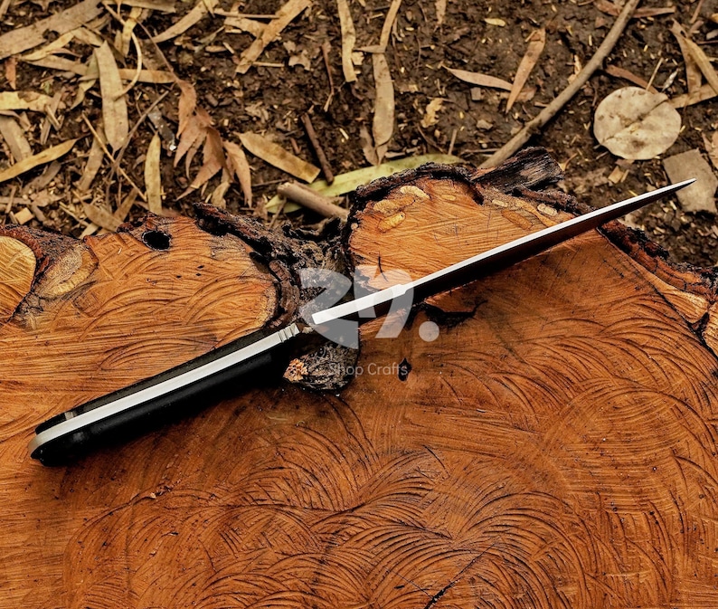 May include: A black-handled knife with a silver blade rests on a textured, brown wooden surface. The knife's blade is sharp and angled, with a visible edge. The handle has a silver stripe. The background includes wood shavings and leaves.