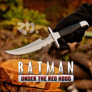 May include: A silver and black knife with a serrated blade and a black handle. The knife is held by a gloved hand. The text "BATMAN UNDER THE RED HOOD" is displayed below the knife.