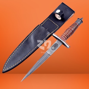 May include: A double-edged dagger with a pointed steel blade, a ribbed brown leather handle, and a metal crossguard marked with the text FSSF. The dagger is accompanied by a black leather sheath featuring yellow stitching and a snap button closure.