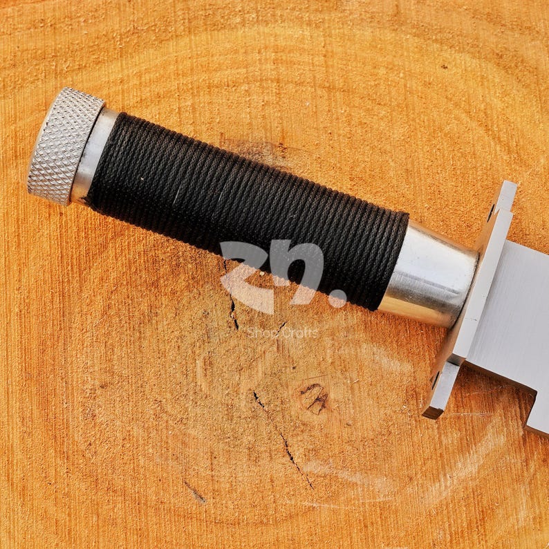 May include: Close-up of a knife handle with a black, textured grip and a silver-colored metal end. The blade is partially visible, made of shiny metal. The knife rests on a wooden surface.