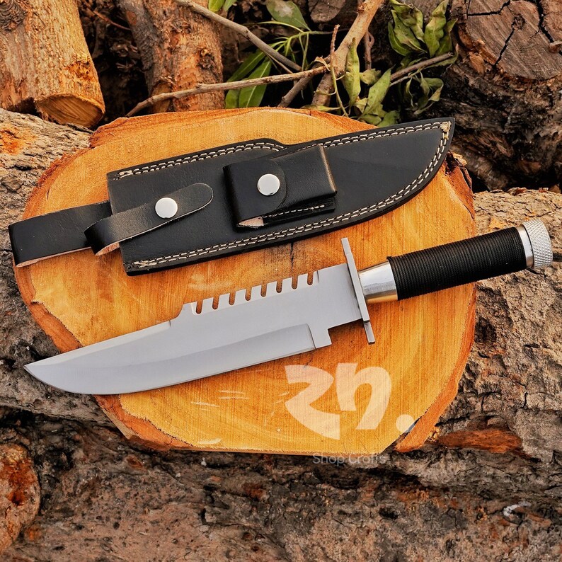 May include: A large, silver-bladed knife with a black handle and a black leather sheath. The knife has a serrated edge and a silver crossguard. The handle is wrapped in black material for grip. The sheath has a belt loop and a snap closure.