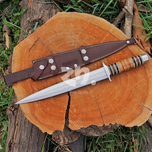 May include: A double-edged fixed blade knife with a pointed silver steel tip, a silver crossguard, and a handle featuring alternating bands of tan, brown, and black material. A brown leather sheath with silver metal snaps lies beside the knife.