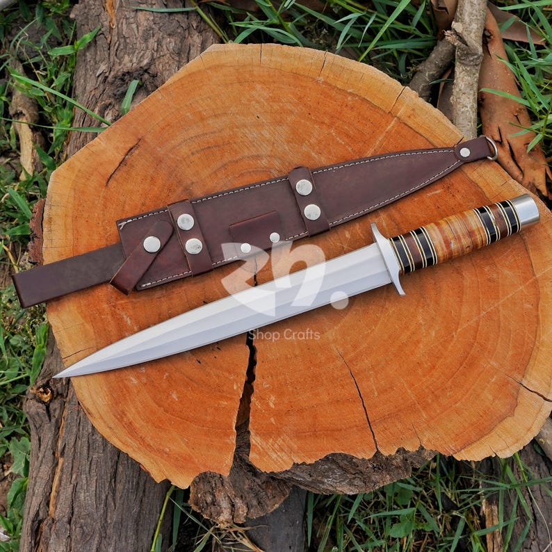 May include: A double-edged fixed blade knife with a pointed silver steel tip, a silver crossguard, and a handle featuring alternating bands of tan, brown, and black material. A brown leather sheath with silver metal snaps lies beside the knife.