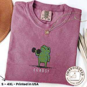 Puede incluir: Camiseta color bayas con un bordado de una rana verde levantando pesas, con el texto "EGADS!". La camiseta tiene cuello redondo y una etiqueta pequeña. Un collar de oro está drapeado sobre la camiseta.
