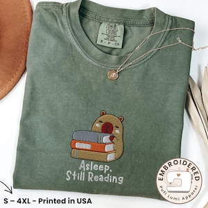 May include: A moss green t-shirt featuring an embroidered graphic of a brown capybara resting its head on a stack of three books. The text below the graphic reads, "Asleep. Still Reading." The shirt has a crew neckline and a Comfort Colors label.