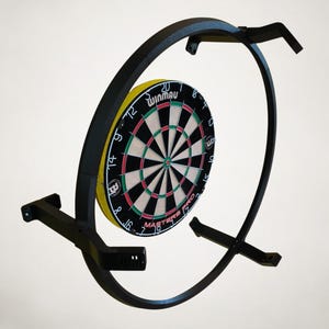 K&ouml;nnte beinhalten: Ein schwarzer Dartscheibenst&auml;nder aus Metall mit einer Winmau Dartscheibe. Die Dartscheibe hat einen gelben Au&szlig;enring, schwarze und gr&uuml;ne Bereiche und wei&szlig;e Zahlen. Der St&auml;nder hat ein gebogenes Design und ist an einer Wand montiert.
