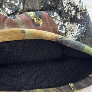 May include: A camouflage beanie hat with a black interior lining. The exterior features a pattern of brown, green, and gray hues, resembling a forest or woodland design. The hat is made of a soft, flexible material.