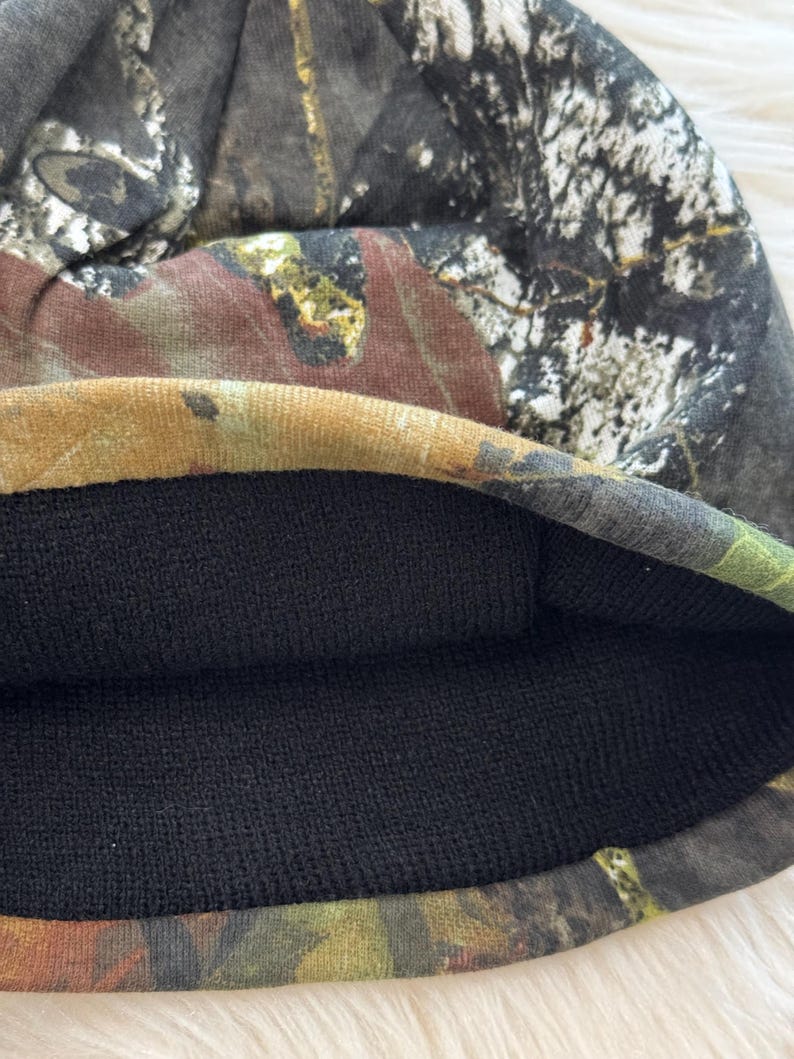 May include: A camouflage beanie hat with a black interior lining. The exterior features a pattern of brown, green, and gray hues, resembling a forest or woodland design. The hat is made of a soft, flexible material.