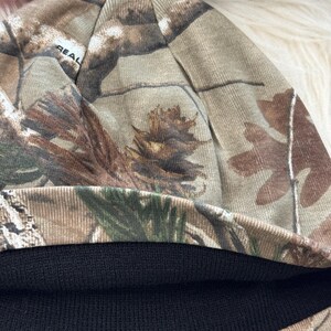 May include: A camouflage beanie hat with a black knit brim. The hat features a realistic forest pattern in shades of brown, green, and tan. The word "REAL" is visible on the side of the hat.