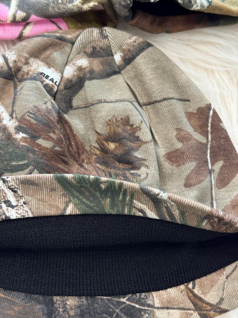 May include: A camouflage beanie hat with a black knit brim. The hat features a realistic forest pattern in shades of brown, green, and tan. The word "REAL" is visible on the side of the hat.