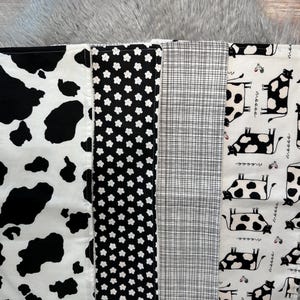 K&ouml;nnte beinhalten: Ein Patchwork-Stoff mit verschiedenen schwarz-wei&szlig;en Mustern. Designs umfassen Kuhflecken, kleine Blumen, ein Karomuster und eine sich wiederholende Kuhillustration. Der Stoff ist wahrscheinlich zum Quilten oder Basteln gedacht.