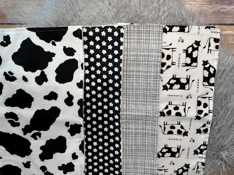 K&ouml;nnte beinhalten: Ein Patchwork-Stoff mit verschiedenen schwarz-wei&szlig;en Mustern. Designs umfassen Kuhflecken, kleine Blumen, ein Karomuster und eine sich wiederholende Kuhillustration. Der Stoff ist wahrscheinlich zum Quilten oder Basteln gedacht.