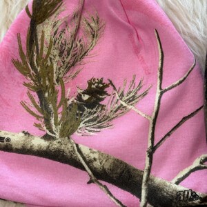 May include: A pink beanie hat with a camouflage pattern featuring branches and foliage in shades of brown and green. The hat is made of a soft, stretchy material and is designed to fit snugly on the head.