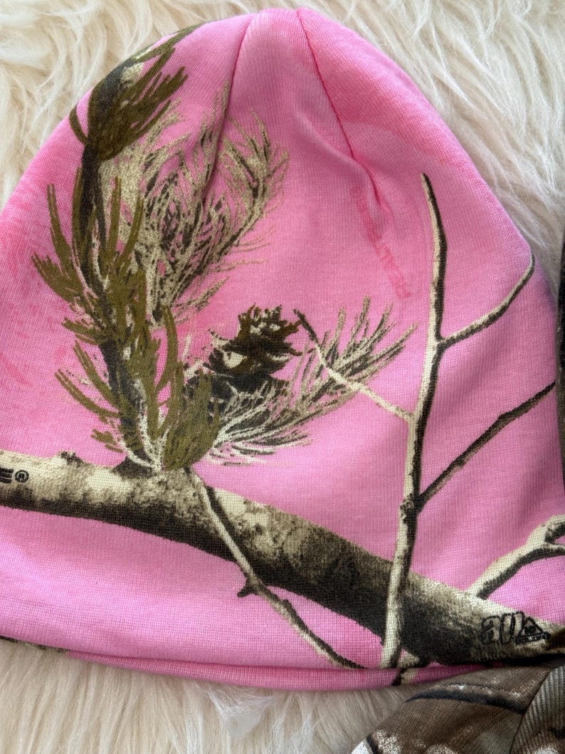 May include: A pink beanie hat with a camouflage pattern featuring branches and foliage in shades of brown and green. The hat is made of a soft, stretchy material and is designed to fit snugly on the head.