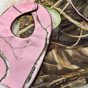 May include: Two baby bibs. One is pink with a Realtree camouflage pattern and the other is brown and green camouflage. The pink bib has a brown trim and the brown and green bib has a neon green trim.