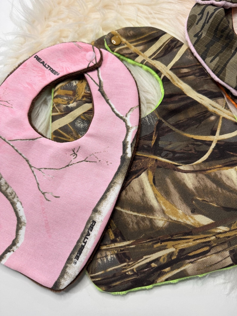 May include: Two baby bibs. One is pink with a Realtree camouflage pattern and the other is brown and green camouflage. The pink bib has a brown trim and the brown and green bib has a neon green trim.
