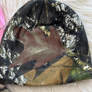 May include: A camouflage beanie hat featuring a brown leaf design. The hat is primarily black, brown, and green, with a woodland camouflage pattern. The beanie is made of a soft material.