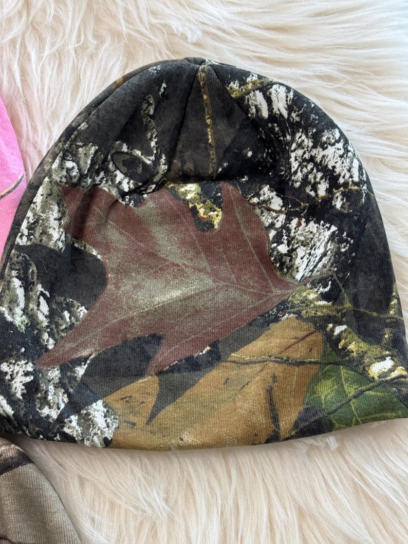 May include: A camouflage beanie hat featuring a brown leaf design. The hat is primarily black, brown, and green, with a woodland camouflage pattern. The beanie is made of a soft material.