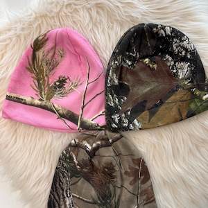 May include: Three beanies with camouflage patterns. One is pink with brown and green branches, another is brown and black with leaves, and the third is brown and green. The beanies are made of a soft material.