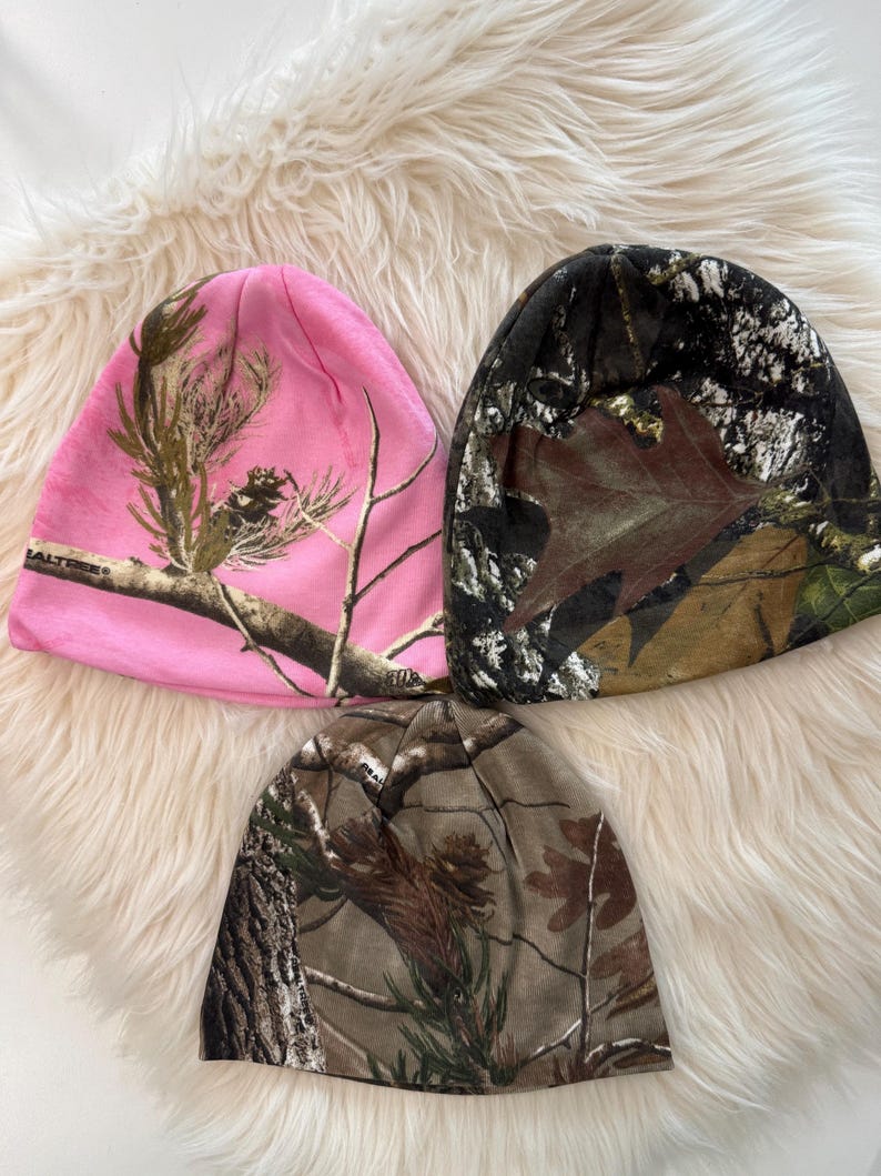 May include: Three beanies with camouflage patterns. One is pink with brown and green branches, another is brown and black with leaves, and the third is brown and green. The beanies are made of a soft material.