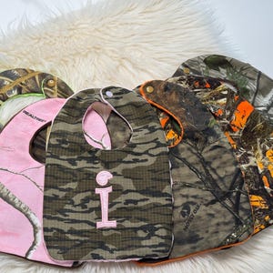 May include: Several baby bibs in camouflage designs. One is pink with a Realtree pattern, and another has a green and brown camouflage with a pink embroidered letter "I". The bibs are made of soft fabric and have a curved shape, suitable for babies.