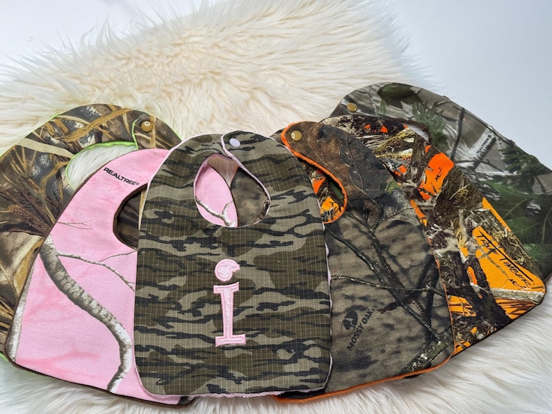 May include: Several baby bibs in camouflage designs. One is pink with a Realtree pattern, and another has a green and brown camouflage with a pink embroidered letter "I". The bibs are made of soft fabric and have a curved shape, suitable for babies.