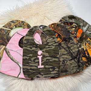 May include: A selection of baby bibs in camouflage designs, including pink and classic green and brown. One bib has a pink letter "i". The bibs are made of fabric and have a curved shape with a neck opening.