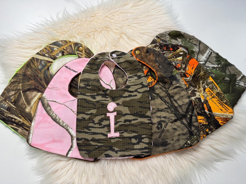 May include: A selection of baby bibs in camouflage designs, including pink and classic green and brown. One bib has a pink letter "i". The bibs are made of fabric and have a curved shape with a neck opening.