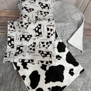 K&ouml;nnte beinhalten: Ein Stapel Babydecken mit Kuh-Motiv. Die Decken haben einen wei&szlig;en Hintergrund mit schwarzen Kuhflecken und Kuh-Illustrationen. Eine Decke hat ein schwarz-wei&szlig;es Karomuster. Die Decken sind weich und gem&uuml;tlich.