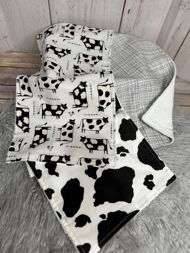 K&ouml;nnte beinhalten: Ein Stapel Babydecken mit Kuh-Motiv. Die Decken haben einen wei&szlig;en Hintergrund mit schwarzen Kuhflecken und Kuh-Illustrationen. Eine Decke hat ein schwarz-wei&szlig;es Karomuster. Die Decken sind weich und gem&uuml;tlich.