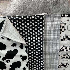 K&ouml;nnte beinhalten: Eine Auswahl an Baby-Spuckt&uuml;chern mit verschiedenen Mustern. Designs umfassen einen Kuhdruck, schwarz-wei&szlig;e Blumen, ein Karomuster und ein wei&szlig;es Tuch mit schwarzen Kuhillustrationen. Die T&uuml;cher sind aus weichem Stoff.