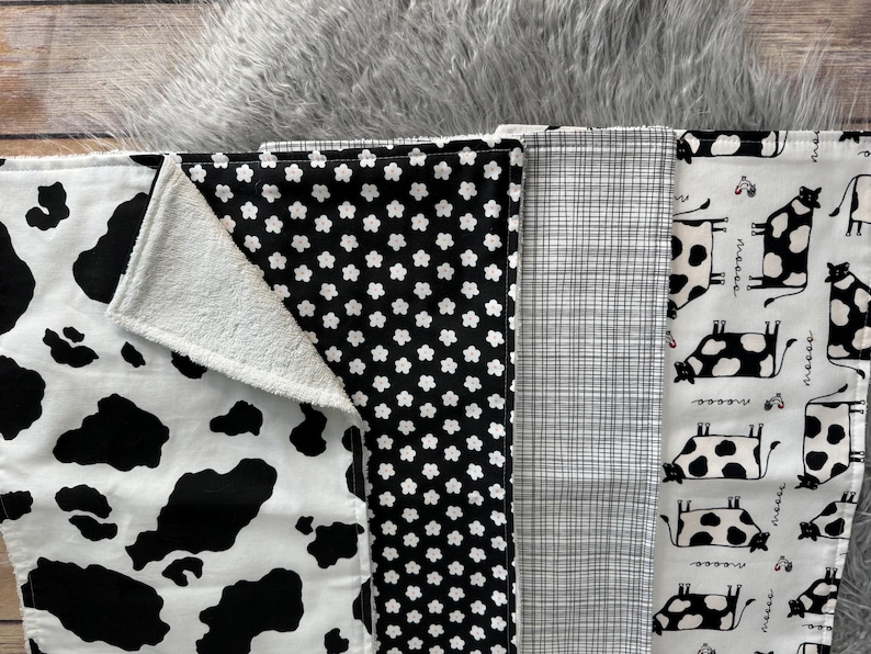 K&ouml;nnte beinhalten: Eine Auswahl an Baby-Spuckt&uuml;chern mit verschiedenen Mustern. Designs umfassen einen Kuhdruck, schwarz-wei&szlig;e Blumen, ein Karomuster und ein wei&szlig;es Tuch mit schwarzen Kuhillustrationen. Die T&uuml;cher sind aus weichem Stoff.