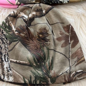 May include: A camouflage beanie hat with a brown, green, and tan pattern. The hat has a "REAL TREE" logo. Another pink camouflage hat is partially visible in the background.