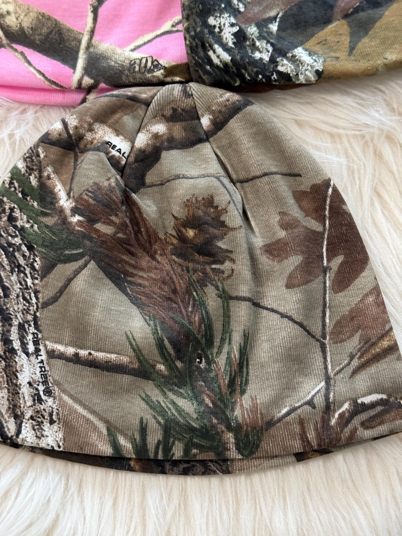 May include: A camouflage beanie hat with a brown, green, and tan pattern. The hat has a "REAL TREE" logo. Another pink camouflage hat is partially visible in the background.